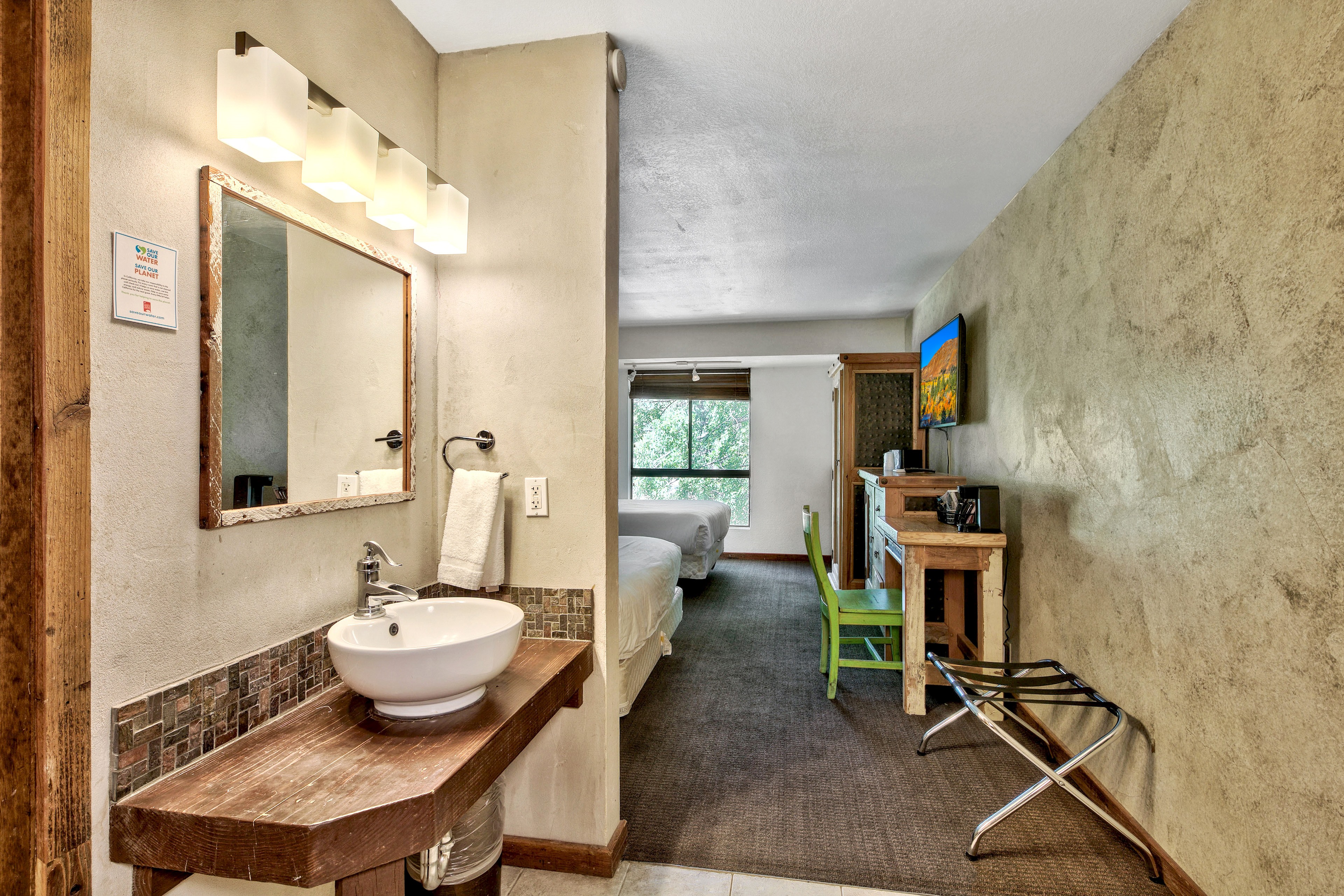 A hotel room with a bathroom sink, mirror, and a bed in the background.