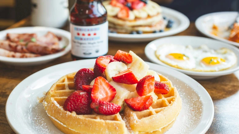 A breakfast spread with a waffle topped with strawberries and bananas, eggs, bacon, and syrup on a wooden table.